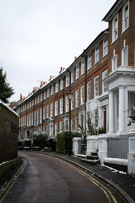 A row of traditional red-brick terraced houses with white window frames and decorative columns at the entrance, situated along a narrow, slightly curved street in a suburban area. The pavement on the right is bordered by low white walls and black metal railings, with some steps leading up to the front doors. The houses feature pitched roofs with small chimney stacks visible against an overcast sky. In the foreground, a black lamppost stands close to the pavement, and there are small leafless trees and bushes next to the houses, some of which are dusted with light snow. The street appears quiet and clean, with a slight accumulation of snow along the edges of the road and pavement, suggesting a recent snowfall. The environment, with its tidy appearance and residential setting, could be indicative of a location where private waste collection services, such as those offered by Waste Removal Twickenham, are used as an alternative to local authority rubbish collection, especially in properties with on-site waste management arrangements.