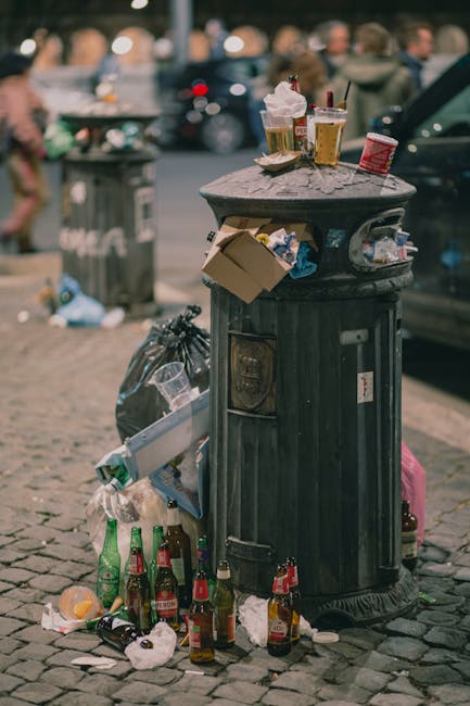 A black, round public trash bin with a domed lid situated on a cobblestone street, heavily surrounded and topped with assorted litter, including several empty glass bottles, some green and others brown, scattered at its base and leaning or stacked around it. On top of the bin, there are multiple food and drink containers, such as a paper coffee cup with a plastic lid, a clear plastic cup filled with a beverage, a small red and white paper container, and a foil-wrapped item, some of which are also placed directly on the lid. The litter spills over onto the cobblestones in front of the bin, with additional items like crumpled paper, plastic bags, and small food wrappers. In the background, blurred figures of pedestrians and parked vehicles are visible, suggesting an urban environment, possibly in a busy street or marketplace area. The scene conveys a moment of communal waste and the importance of private or independent rubbish collection services that Waste Removal Twickenham may provide for maintaining cleaner public spaces.
