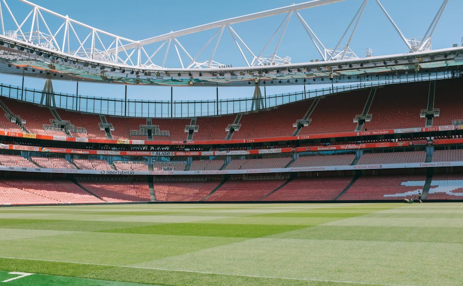 An empty sports stadium with a large, well-maintained grass pitch in the foreground, featuring alternating light and dark green stripes. Surrounding the pitch are multiple tiers of red and pink stadium seats, with some sections having visible white patterns or markings. The seating area includes advertising banners and digital screens, with the one in the middle displaying the website 'Arsenal.com.' Above the seats, a continuous concrete or metal structure supports the upper tiers, and the stadium is partially covered by a curved, white steel framework that forms an open, arching roof structure, allowing natural daylight to illuminate the scene. The sky above is clear and blue, contributing to a bright, well-lit atmosphere. The overall scene is neutral and professional, focusing on the architectural and environmental features of the stadium, with no visible signage or activity, highlighting the space as suitable for potential waste removal and cleaning services that could be required after events.