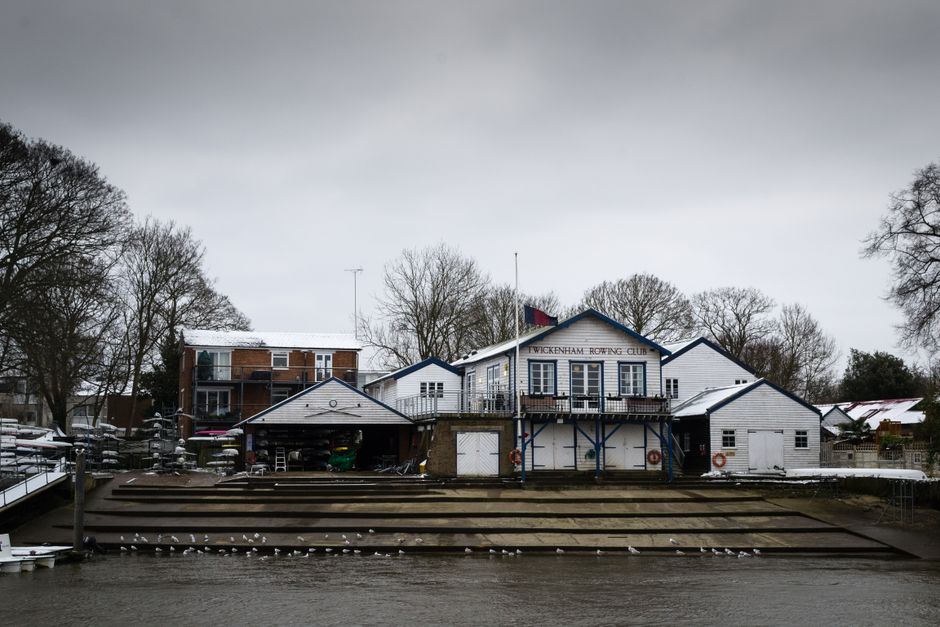 A view of the Twickenham Rowing Club building positioned along the River Thames, featuring a white wooden exterior with blue trim and a flagpole extending from the front. The structure is situated on a concrete shoreline with tiered steps leading down to the water, where a flock of seagulls is perched on the lower steps. Surrounding the building are leafless trees, indicating a winter season, and the sky overhead is overcast with gray clouds. To the left, there are additional boats and equipment stored on the riverbank, and behind the club, residential buildings with brick and timber exteriors are visible. The scene is captured during daylight hours under subdued natural light, reflecting a calm, quiet riverside environment suitable for outdoor activities and club operations, often requiring occasional waste clearance or rubbish removal services by providers like Waste Removal Twickenham.
