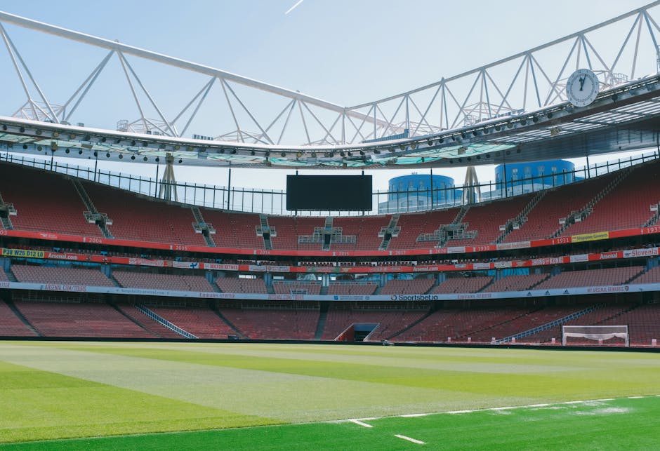 An empty sports stadium with a large, well-maintained grass pitch in the foreground, featuring alternating light and dark green stripes. Surrounding the pitch are multiple tiers of red and pink stadium seats, with some sections having visible white patterns or markings. The seating area includes advertising banners and digital screens, with the one in the middle displaying the website 'Arsenal.com.' Above the seats, a continuous concrete or metal structure supports the upper tiers, and the stadium is partially covered by a curved, white steel framework that forms an open, arching roof structure, allowing natural daylight to illuminate the scene. The sky above is clear and blue, contributing to a bright, well-lit atmosphere. The overall scene is neutral and professional, focusing on the architectural and environmental features of the stadium, with no visible signage or activity, highlighting the space as suitable for potential waste removal and cleaning services that could be required after events.