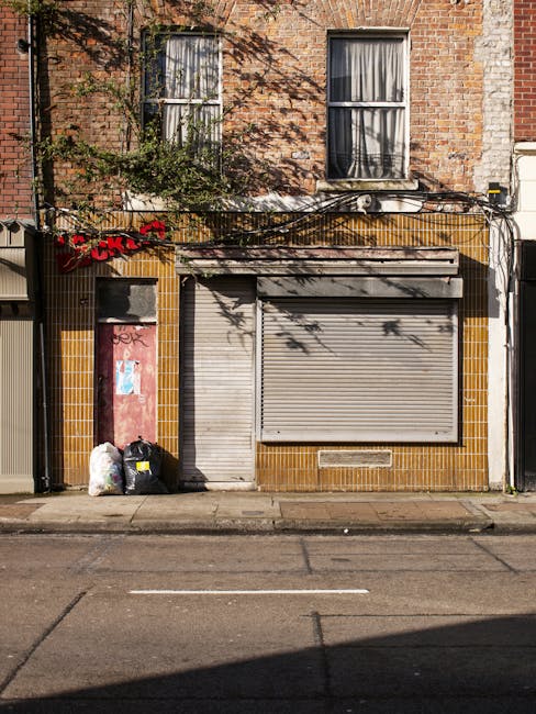 The image depicts a small commercial premises on a city street with a closed metal roller shutter door painted in light grey, occupying the central part of the building's ground floor. To the left of the shutter, there is a narrow, red-painted door with some graffiti, and two black garbage bags and a white plastic bag are placed on the pavement beside it, indicating waste awaiting collection. The building's façade is clad in yellow-brown tiles surrounding the shop entrance, contrasting with the brickwork above, which features two windows on the upper floor with white frames and partially drawn curtains. A sign with red lettering appears to be partially obscured, hanging just above the tiled section. Overhead, a tangle of black electrical wires runs across the façade, casting shadows on the brickwork, and some greenery from a tree branches out toward the upper windows. The overall scene reflects an urban environment where waste removal services, such as those provided by Waste Removal Twickenham, are involved in managing waste materials likely from local shops or small commercial units, emphasizing the importance of private rubbish collection in city streets.