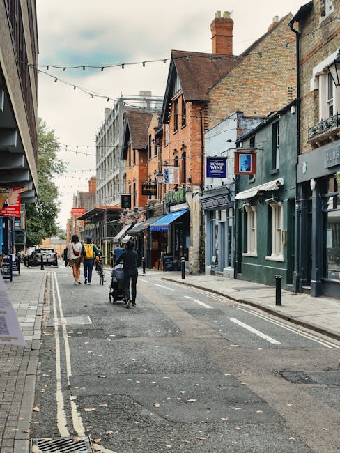 A street scene showing a pedestrian walkway lined with small shops and businesses housed in older brick buildings with varied architectural styles and colors, including green and blue facades. The pavement is paved with small, rectangular stones, and the street has white painted double lines along the edge. Overhead, there are string lights suspended across the street, suggesting a lively, commercial atmosphere. In the foreground, a person pushing a stroller and three other pedestrians are visible walking along the street, which is relatively quiet with minimal vehicle traffic. To the left, a red sign partially visible appears to belonged to a retail shop, while on the right, signs indicate different storefronts, including one advertising wine. The background features more buildings extending along the street, some with additional signage, and trees visible in the distance. The scene likely depicts an area designated for private or independent shopping, related to a rubbish removal service assisting with local waste management in such urban commercial environments, supporting the safe and efficient disposal of waste generated by shops and visitors.