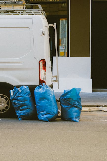 The image depicts the rear side of a white commercial van parked on a street outside a building entrance. Three large blue rubbish bags, made of crinkled plastic material, are placed on the pavement directly behind the vehicle, indicating a collection or waste disposal activity. The bags are filled, with visible creases and slight bulges, suggesting they contain household or light waste. The van's rear red and clear light cluster is visible on the left side, along with part of the black wheel and tire. In the background, to the right, a portion of the building's exterior wall and step is visible, with a glass door or window reflecting some interior light. The scene appears to be in an urban or suburban environment, typical of a private rubbish collection or on-site waste clearance service offered by Waste Removal Twickenham, supporting alternative waste handling options outside of local authority rubbish collection.