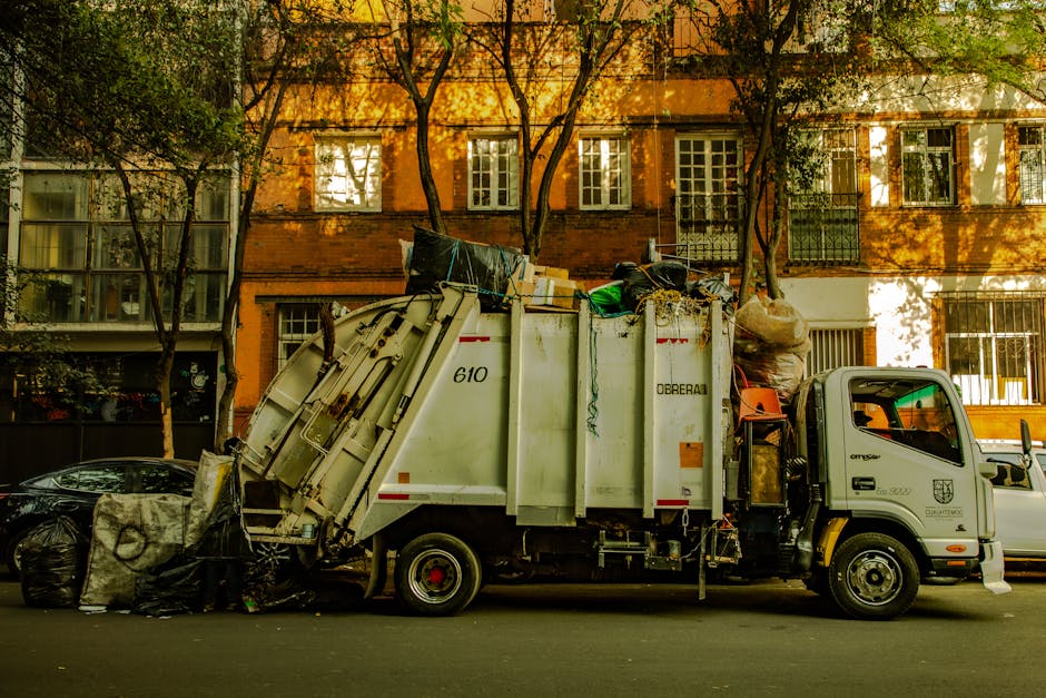 A white waste collection truck operated by Waste Removal Twickenham is parked on a city street during daylight hours, loaded with various types of rubbish and waste materials. The truck's body features a large, enclosed compartment with visible markings and identification numbers, and its rear section is tilted for unloading. The waste includes black and green plastic bags, cardboard boxes, and other miscellaneous refuse, some stacked on top of each other and held in place by securing straps. The vehicle is positioned adjacent to parked cars, with a row of multi-story brick residential buildings in the background, illuminated by warm sunlight filtering through the trees. The surroundings show a typical urban environment with trees providing partial shade, and the street appears quiet with no visible pedestrians. This scene illustrates a private waste collection service, typical of independent rubbish removal operations aimed at on-site clearance in residential areas.